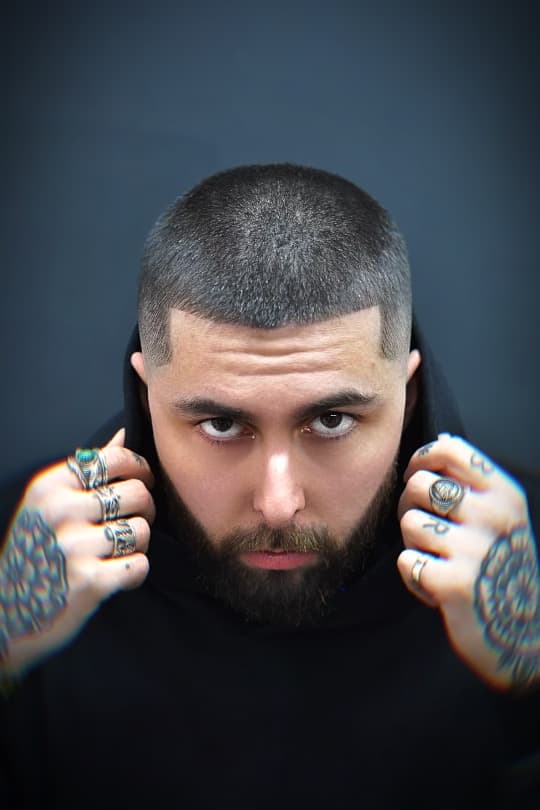 Man with buzz cut and beard stares intensely, holding his hood with tattooed hands.